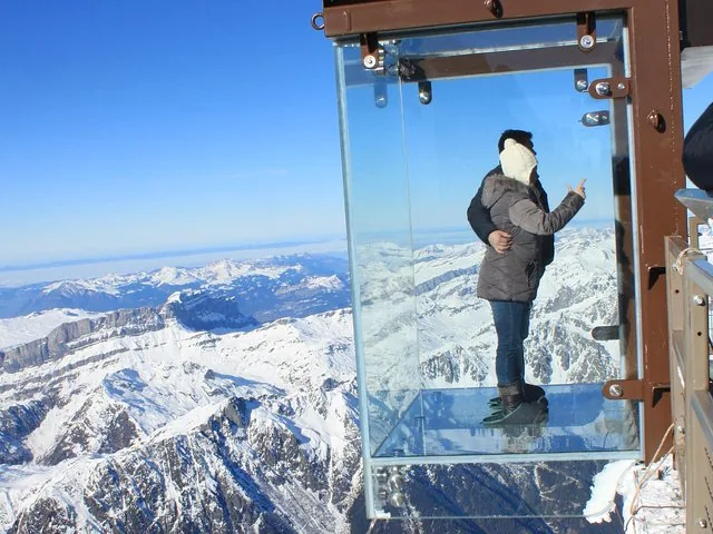 Couple inside a glass viewing box suspended over snowy mountains